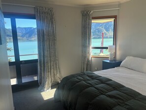 A bedroom with a bed, a nightstand, a lamp, and a window with a view of a lake and mountains. - Cozy retreat, accessible by road and water, with amazing views! (Moetapu Bay)