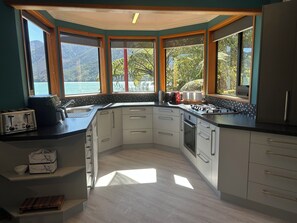 A modern kitchen with a corner island, built-in appliances, and large windows offering a view of the outdoors. - Cozy retreat, accessible by road and water, with amazing views! (Moetapu Bay)