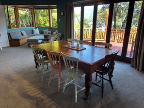 A dining area with a wooden table and chairs, a sofa, and a view of a wooden deck through large windows. - Cozy retreat, accessible by road and water, with amazing views! (Moetapu Bay)