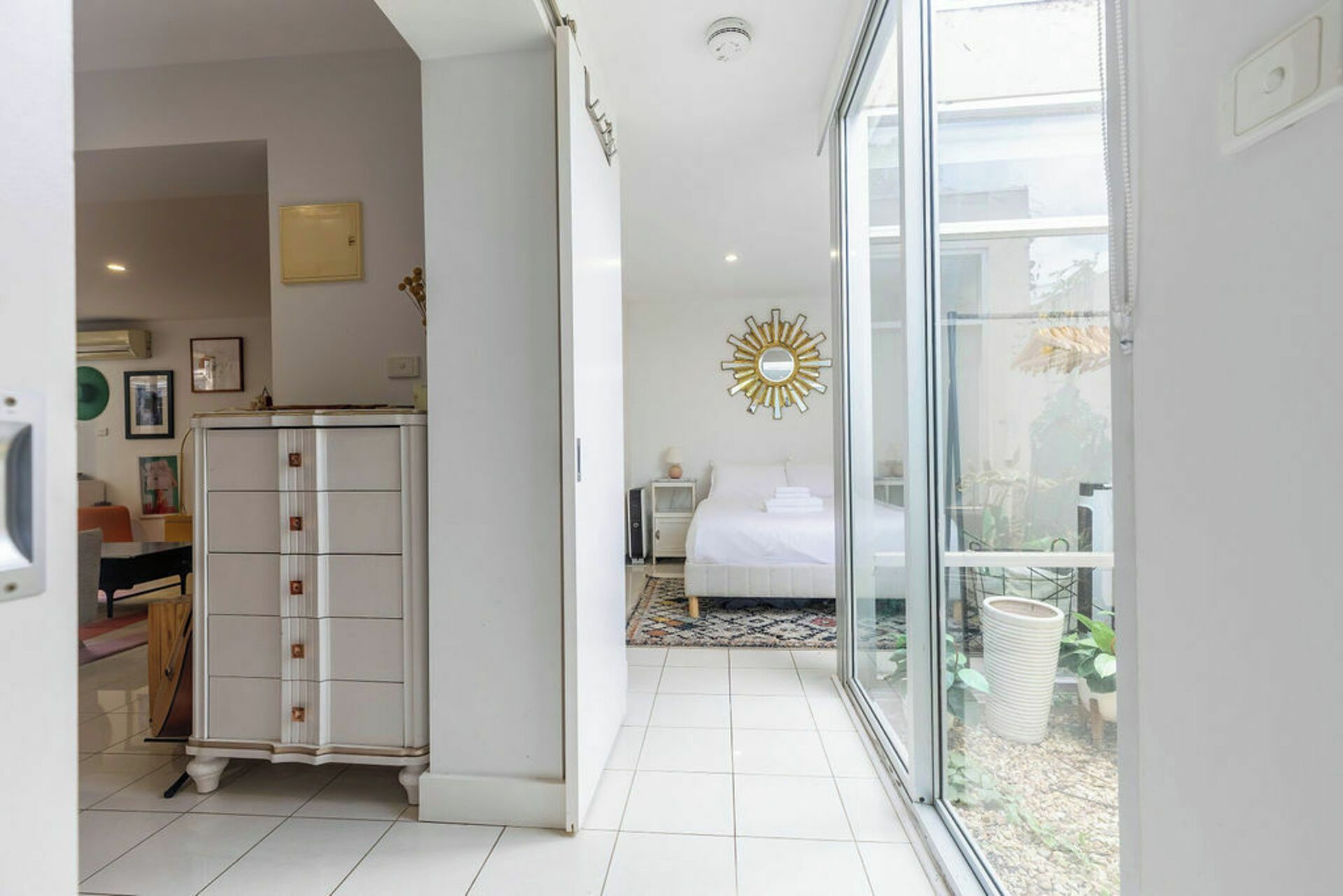 A hotel room with a bed, a nightstand, a dresser, and a large window offering a view of the outdoors.