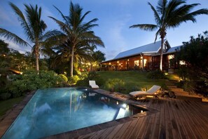 A pool area with lounge chairs, a wooden deck, and a house with a sloped roof and large windows. - Villa Maido Luxe (Saint-Paul)