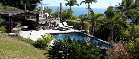 A pool area with lounge chairs, a wooden deck, and a view of the ocean.
