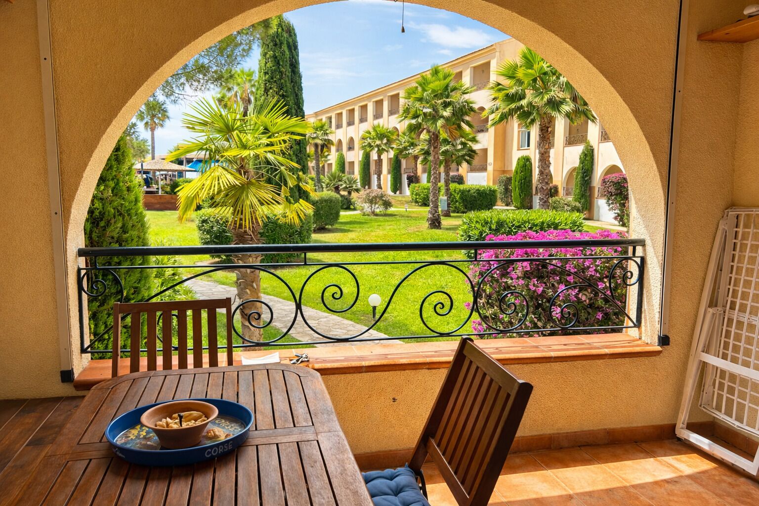 Ein Balkon mit Holztisch, Stuhl und Blick auf einen Innenhof mit Palmen und einem Gebäude.