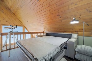 Un dormitorio con techo de madera, un telescopio, una cama con manta, una lámpara de noche y una ventana.