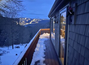Una terraza de madera con acumulación de nieve, un ventanal de vidrio y vistas a montañas nevadas.