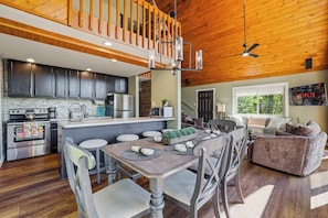 A modern kitchen with stainless steel appliances, a dining area with a wooden table and chairs, a cozy living room with a sofa, and a wooden ceiling with a ceiling fan.