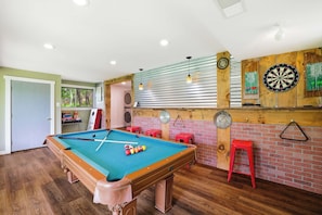 A pool table with balls, a dartboard, and stools in a room with wooden floors and brick walls.