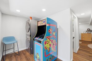 Une petite pièce comprenant un lave-linge, une borne d’arcade bleue, une chaise bleue et une commode en bois.