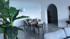 Un coin repas avec une table dressée pour quatre personnes, entourée de chaises. Une grande plante verte se trouve à gauche et un canapé est visible au premier plan. Une porte en arc donne accès à une autre pièce.