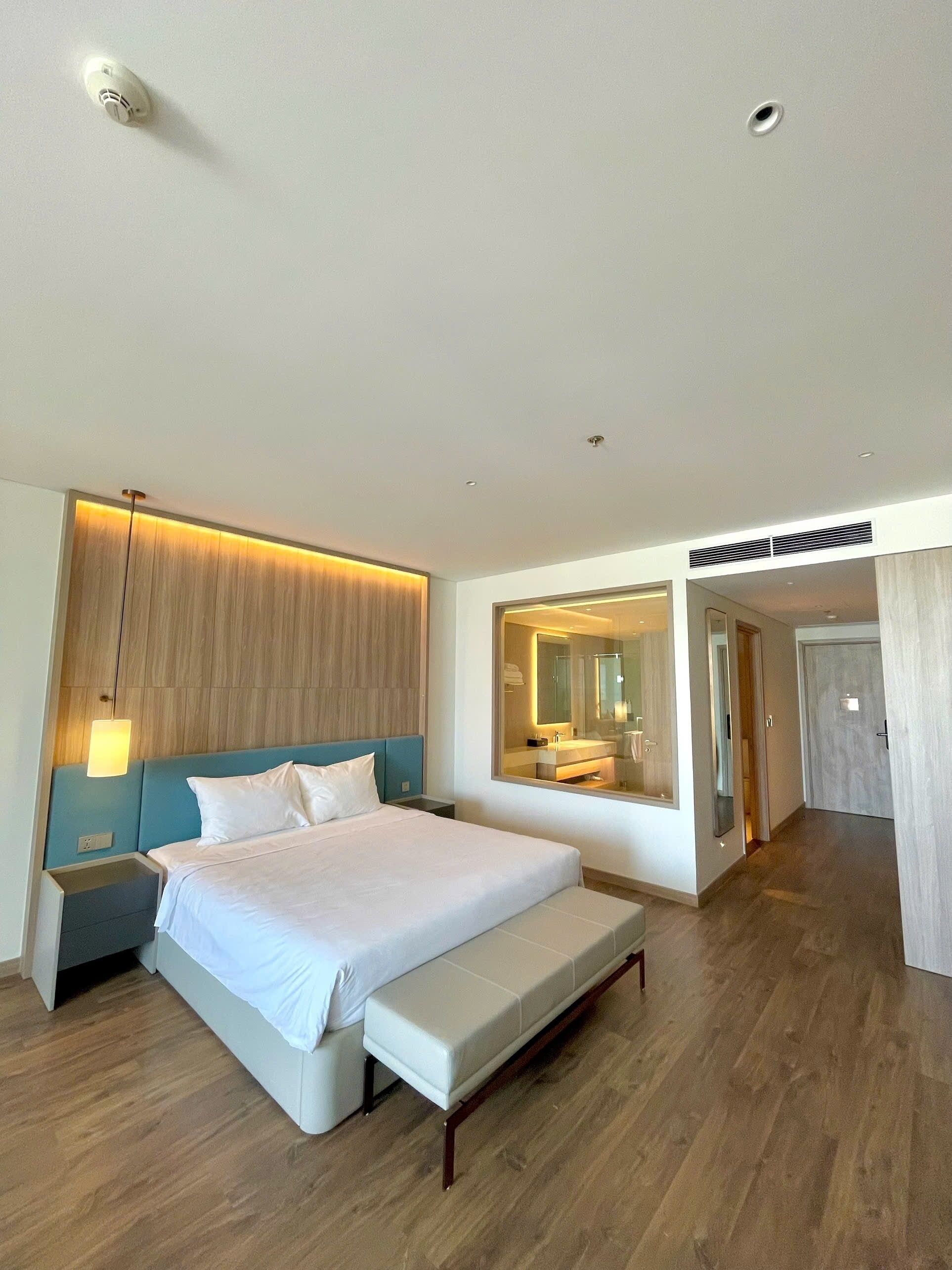A modern hotel room with a large bed, a bedside table, a bench, a mirror, and a wooden panel wall.