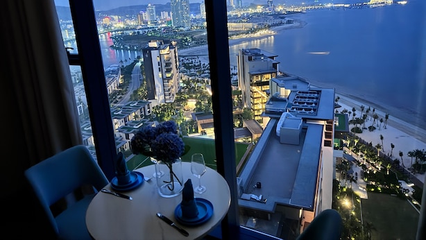 A table set for dining with a vase of flowers, overlooking a coastal cityscape with buildings and a beach. - Alacarte Seaview Halong by HaVan (Bai Chay)