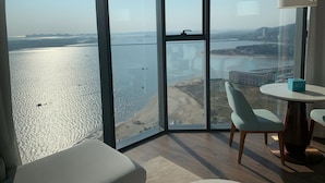 A modern room with a large floor-to-ceiling window offering a view of a beach and cityscape. - Alacarte Seaview Halong by HaVan (Bai Chay)