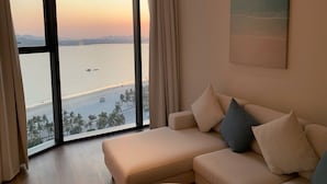 A living room with a large window overlooking a body of water, a beige sofa, and a painting on the wall. - Alacarte Seaview Halong by HaVan (Bai Chay)