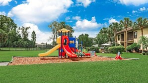 Ein Spielplatz mit Rutsche und Klettergerüsten, umgeben von einem gepflegten Rasen und Palmen.