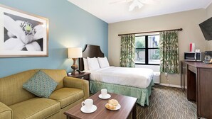 Ein Hotelzimmer mit Bett, Sofa, Schreibtisch mit Computer, Fenster mit Vorhängen und einem Bild an der Wand.