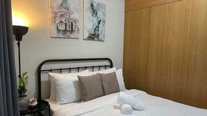 Una cama bien hecha con sábanas blancas y una toalla enrollada, un cabecero de metal negro, y dos cuadros de estructuras arquitectónicas en la pared.