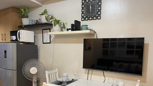 Una cocina compacta con refrigerador, microondas y una mesa para dos personas con sillas. Hay un televisor montado en la pared y un reloj sobre la encimera.