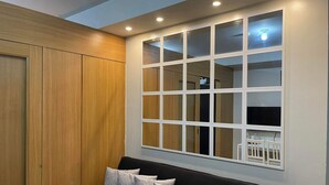 Una habitación con un sofá negro, tres almohadas grises, un armario de madera y un ventanal de varios cristales.