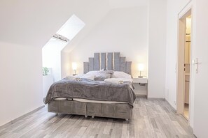 Ein modernes Schlafzimmer mit Schrägen, einem Bett mit grauem Kopfteil, Nachttischlampen und Blick auf einen Flur.