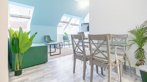 Un soggiorno moderno con un divano verde, un tavolo da pranzo in vetro e due sedie. Sono presenti due piante in vaso, una a sinistra e una a destra. La stanza ha una parete d'accento azzurra e un'ampia finestra che offre luce naturale.