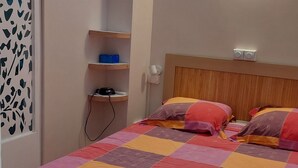 Een slaapkamer met een bed, een raam, een plank met spullen en een wandlamp.