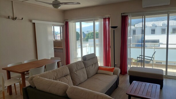 Een woonkamer met een bank, een houten tafel en een eethoek. Er is uitzicht op een gebouw en een balkon met rode gordijnen.