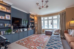 A living room with a large flat-screen TV, a patterned rug, a sofa, and a bookshelf filled with books and decorative items. - Beautiful 5BDR 3Bath Home in Barnes (Greater London)
