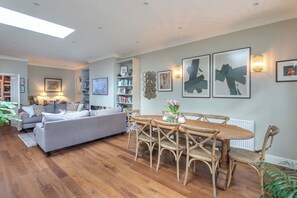 A living room with a sofa, a dining table with chairs, a bookshelf, and wall art. - Beautiful 5BDR 3Bath Home in Barnes (Greater London)