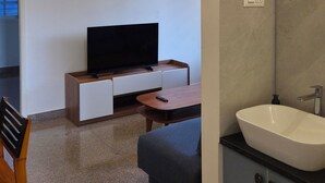 Ein modernes Badezimmer mit Waschbecken, Spiegel und Waschtisch. Im angrenzenden Raum befindet sich ein Fernseher, ein Holzkabinett und ein Sessel.
