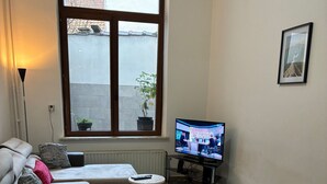 Ein Wohnzimmer mit einer Couch, einem kleinen runden Tisch, einem Fernseher und einem großen Fenster mit Blick auf ein Gebäude und einen Baum.