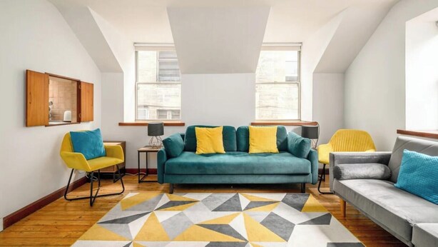 En moderne loftlejlighed med en tealfarvet sofa, gule lænestole og en måtte med geometrisk mønster.