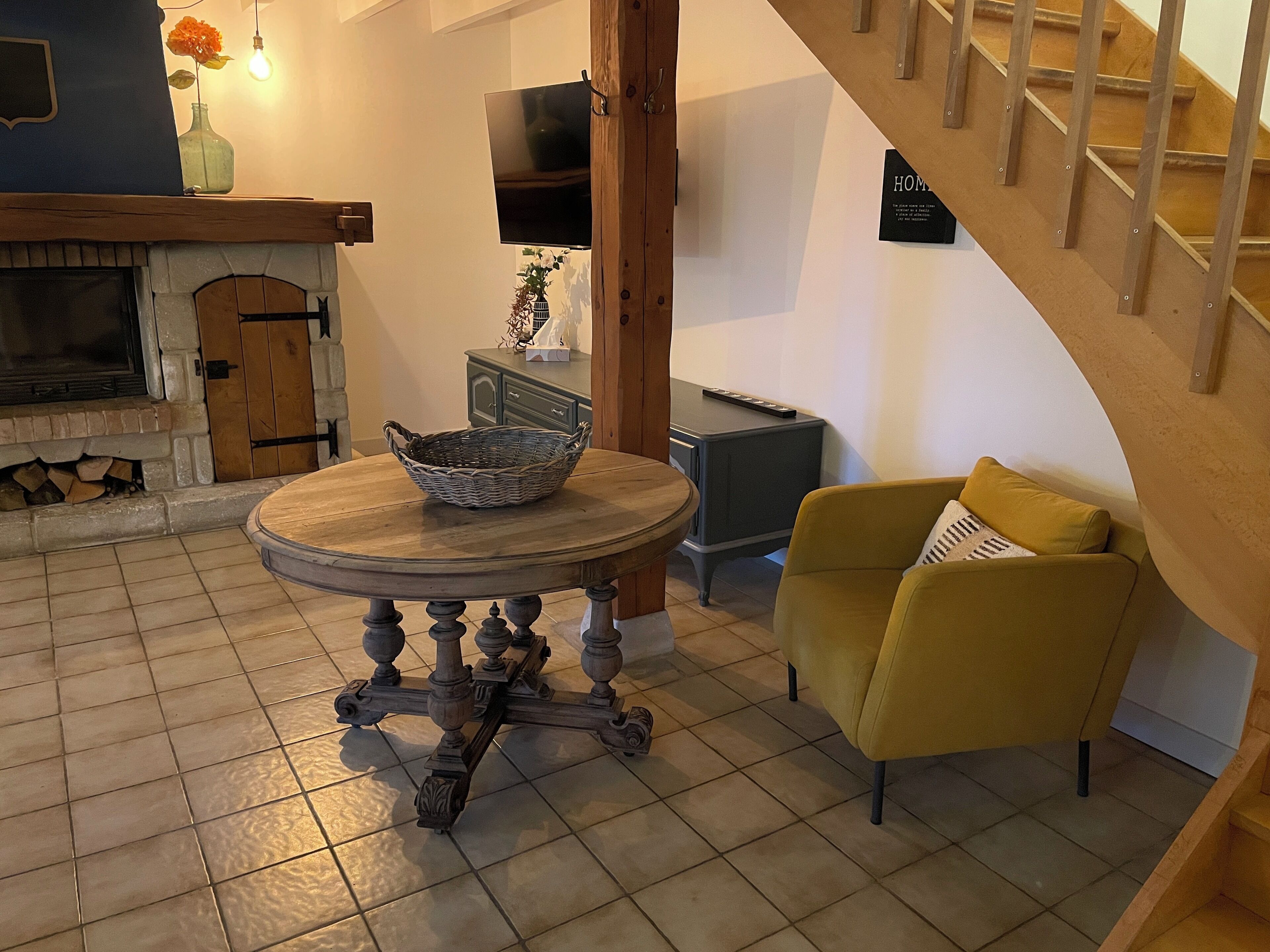 Un salon chaleureux avec une cheminée, une table ronde en bois sur laquelle est posé un panier, un fauteuil jaune et un escalier.