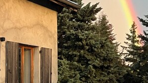 Un edificio con ventana y contraventanas, un árbol al fondo y un arcoíris en el cielo.