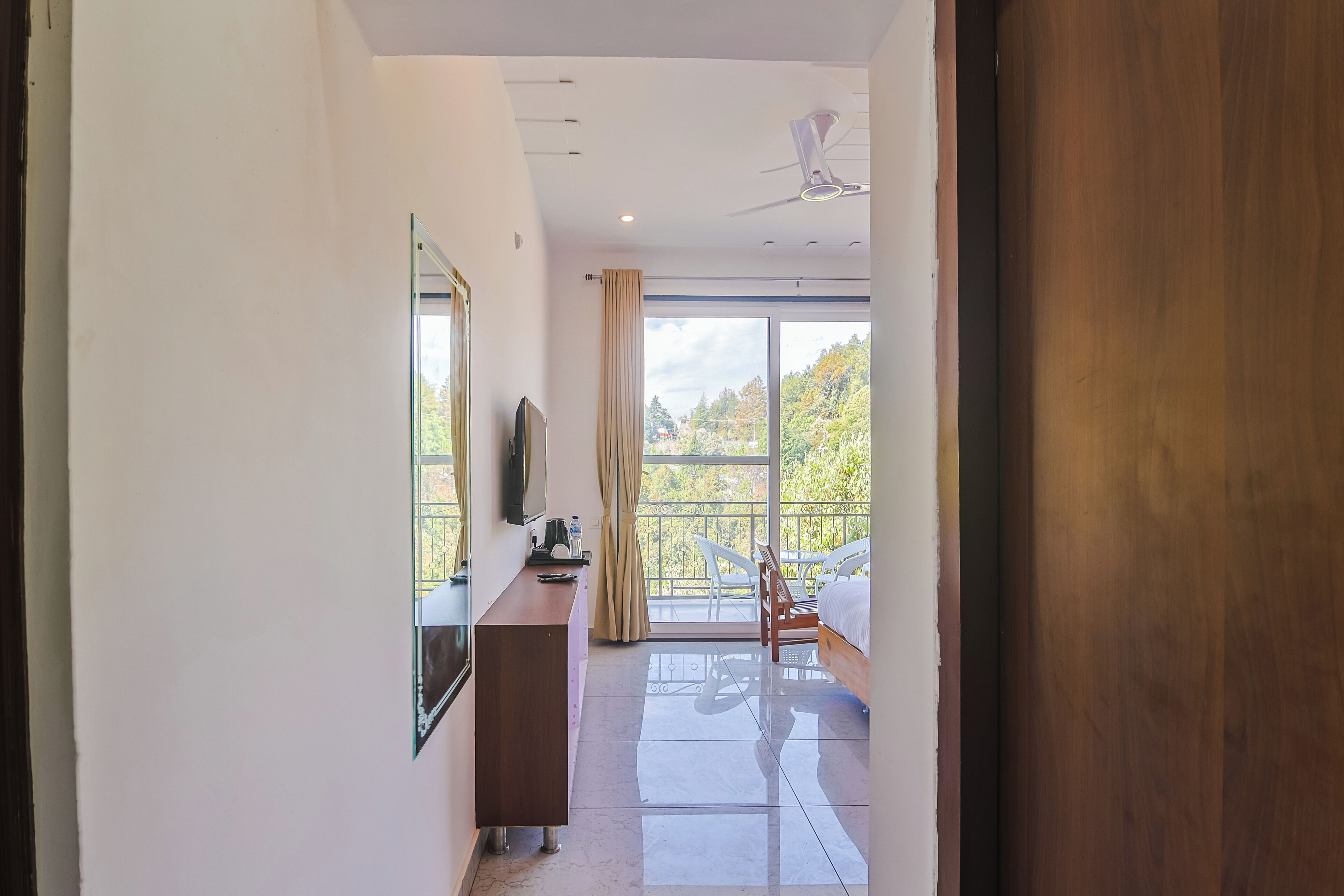 A hotel room with a bed, a desk, a TV, and a balcony with a view of trees.