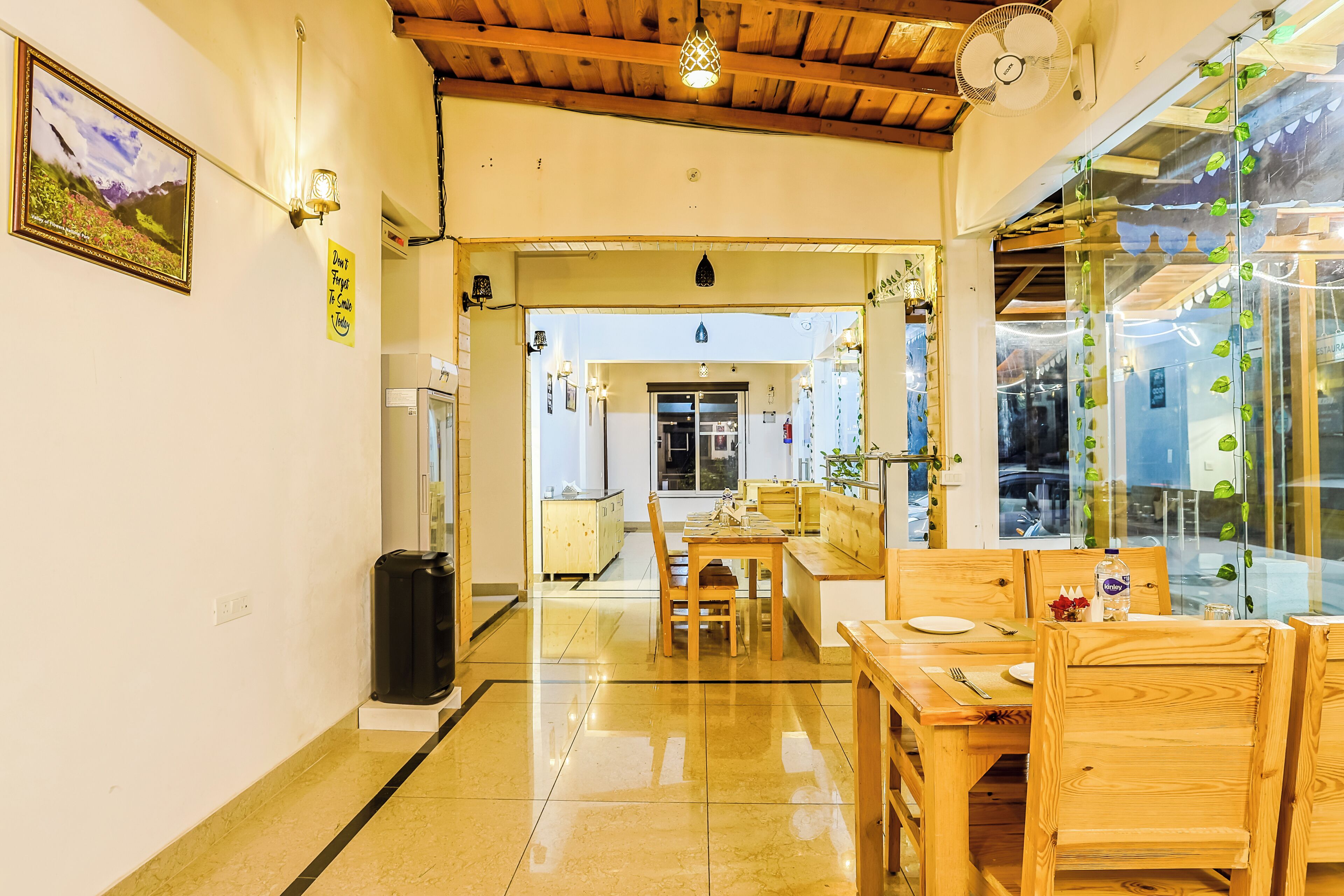 A well-lit dining area with wooden furniture, a glass wall with plants, and a view into another room.