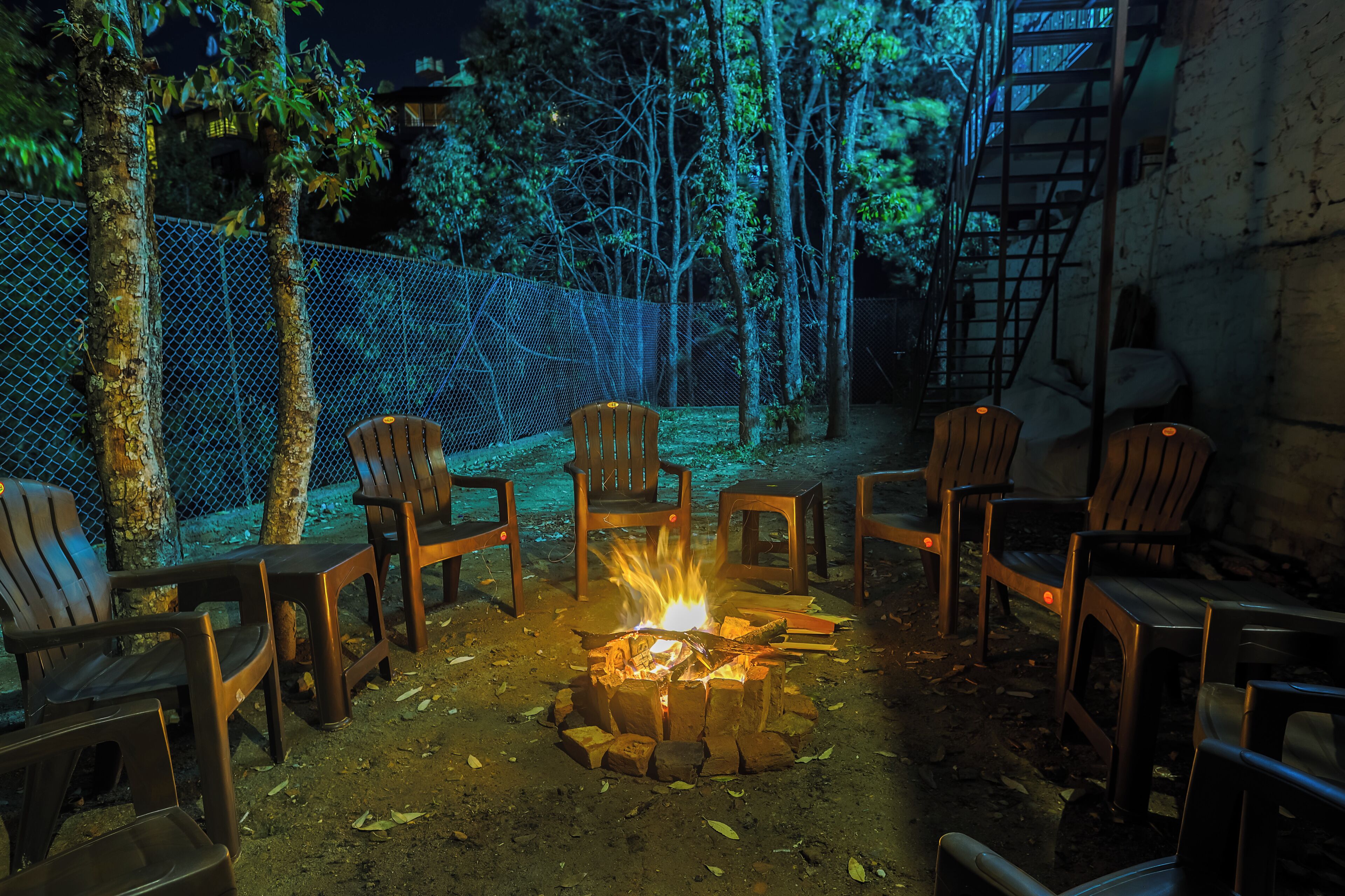 A fire pit surrounded by chairs in an outdoor setting with a chain-link fence and trees in the background.