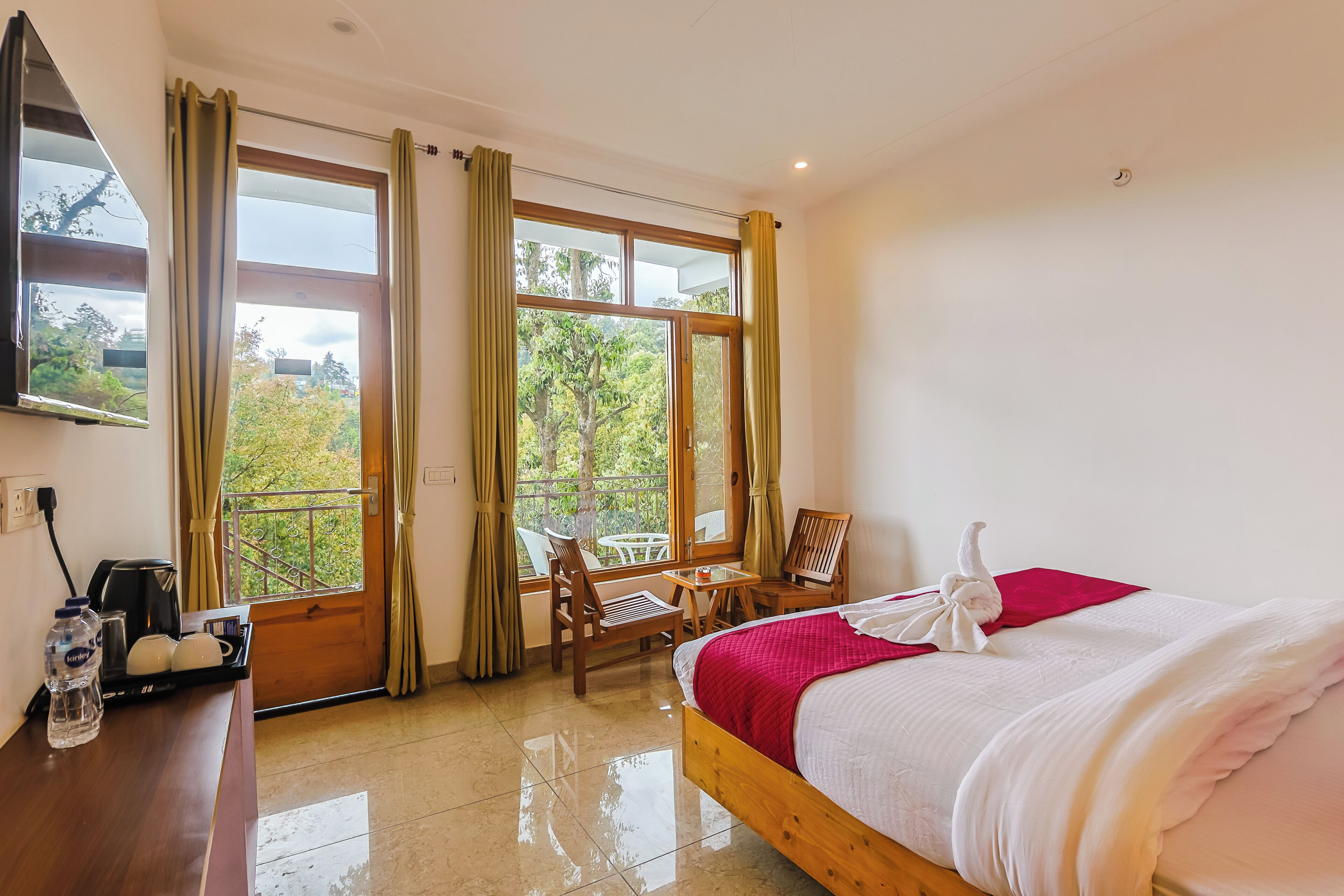 Habitación de hotel con cama, escritorio con tetera, una silla, una mesa y vistas a zonas verdes a través de amplios ventanales.