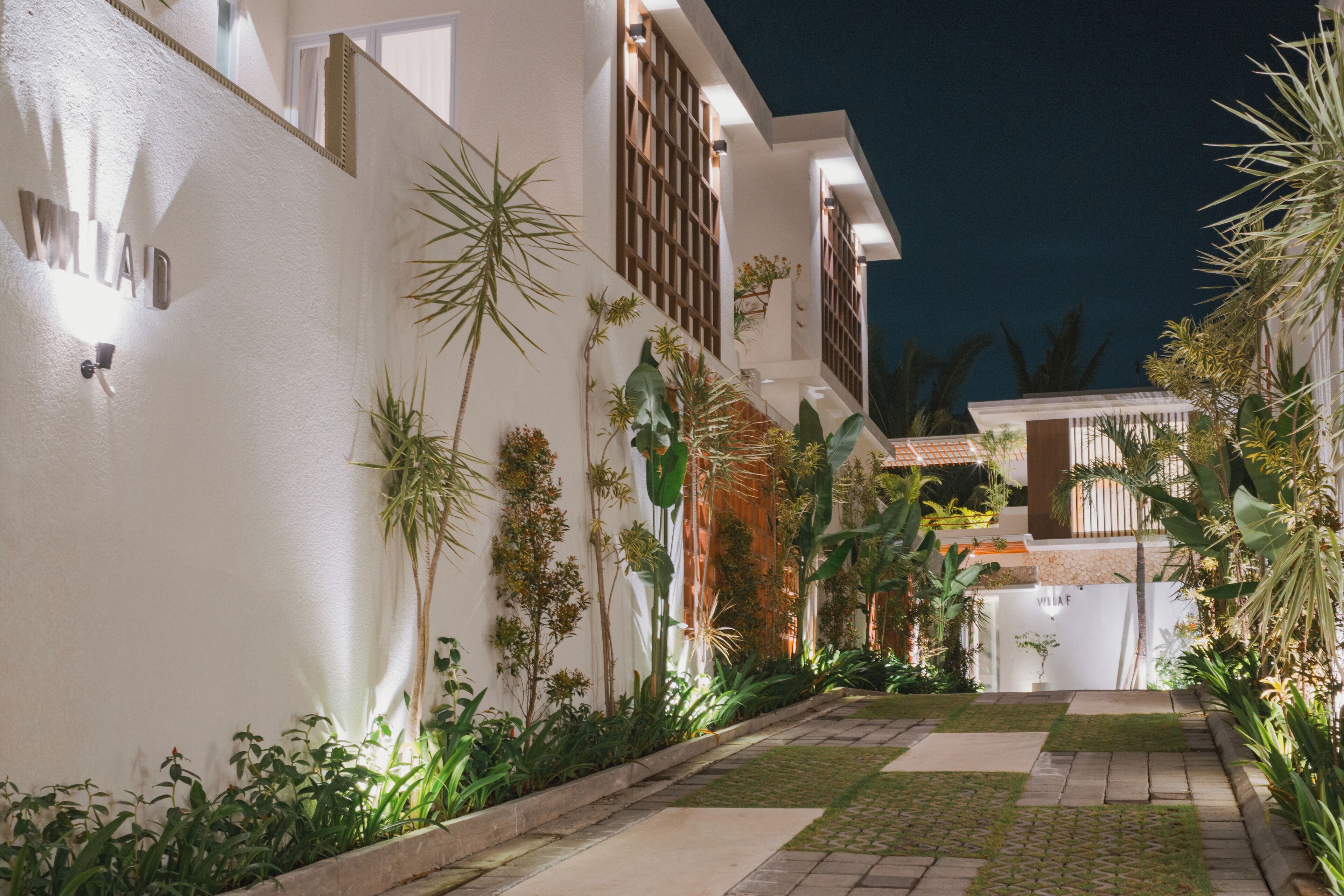 Un edificio moderno con fachada blanca, amplios ventanales y un jardín cuidado con diversas plantas y un sendero pavimentado.
