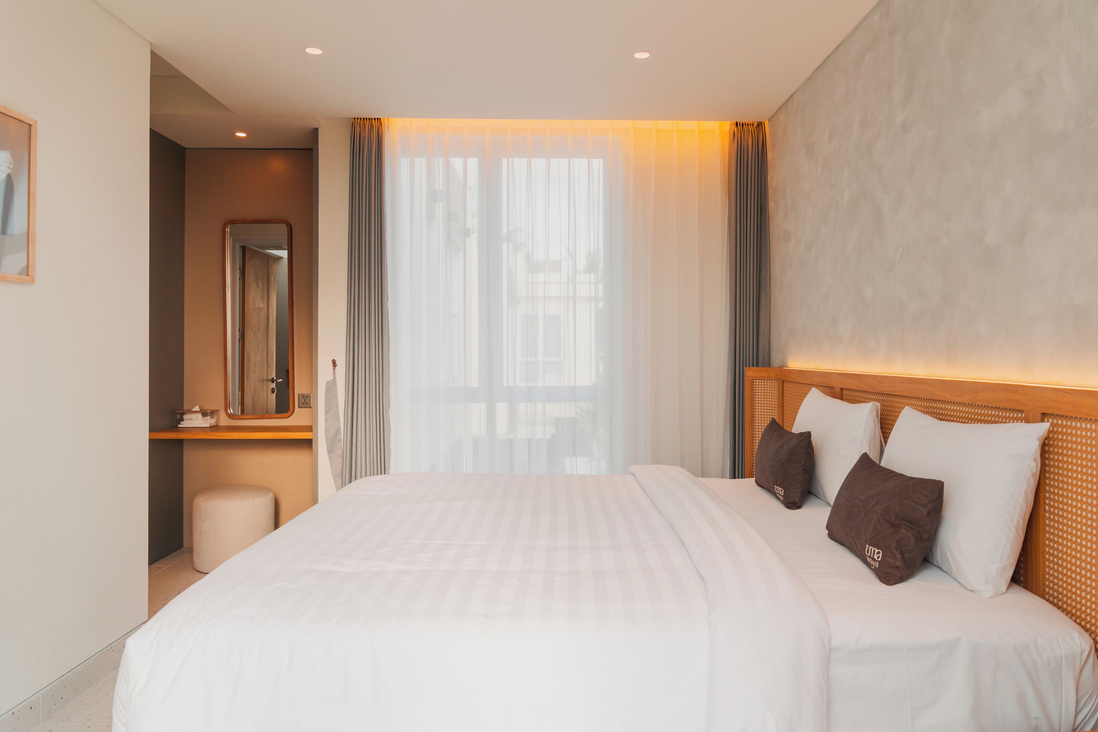A hotel room with a large bed, a wooden headboard, a mirror, and a window with sheer curtains.