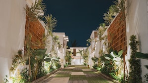 A narrow pathway flanked by white buildings with wooden lattice windows, leading to a central area with a bench and a table. - Uma Avaya Villas Canggu (Canggu)
