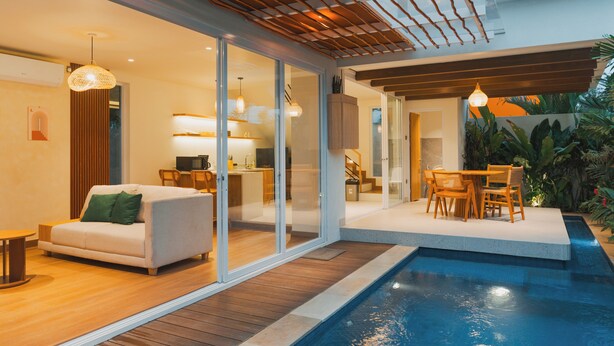A modern indoor pool area with a wooden deck, a white sofa, and a dining set. - Uma Avaya Villas Canggu (Canggu)