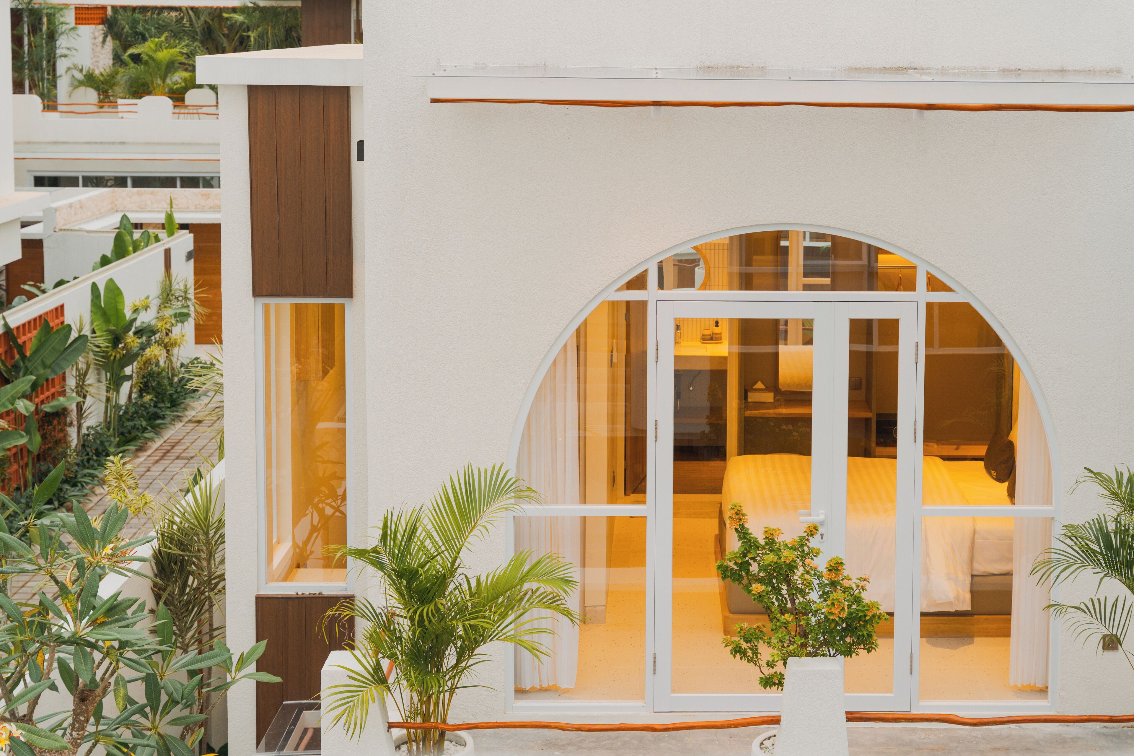 A modern hotel with a large arched window, a white exterior, and a balcony with plants.