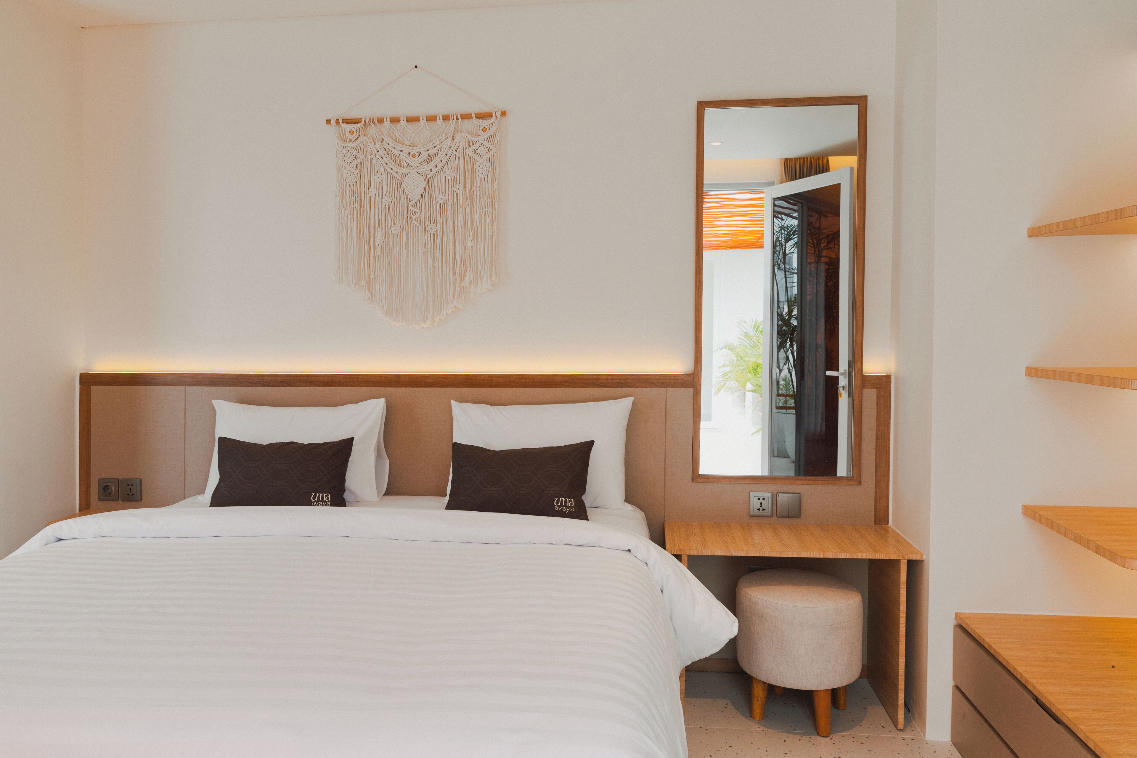 Een moderne slaapkamer met een groot bed, een houten nachtkastje, een rond poefje, een macramé wanddecoratie en een spiegel die uitzicht op de natuur weerkaatst.