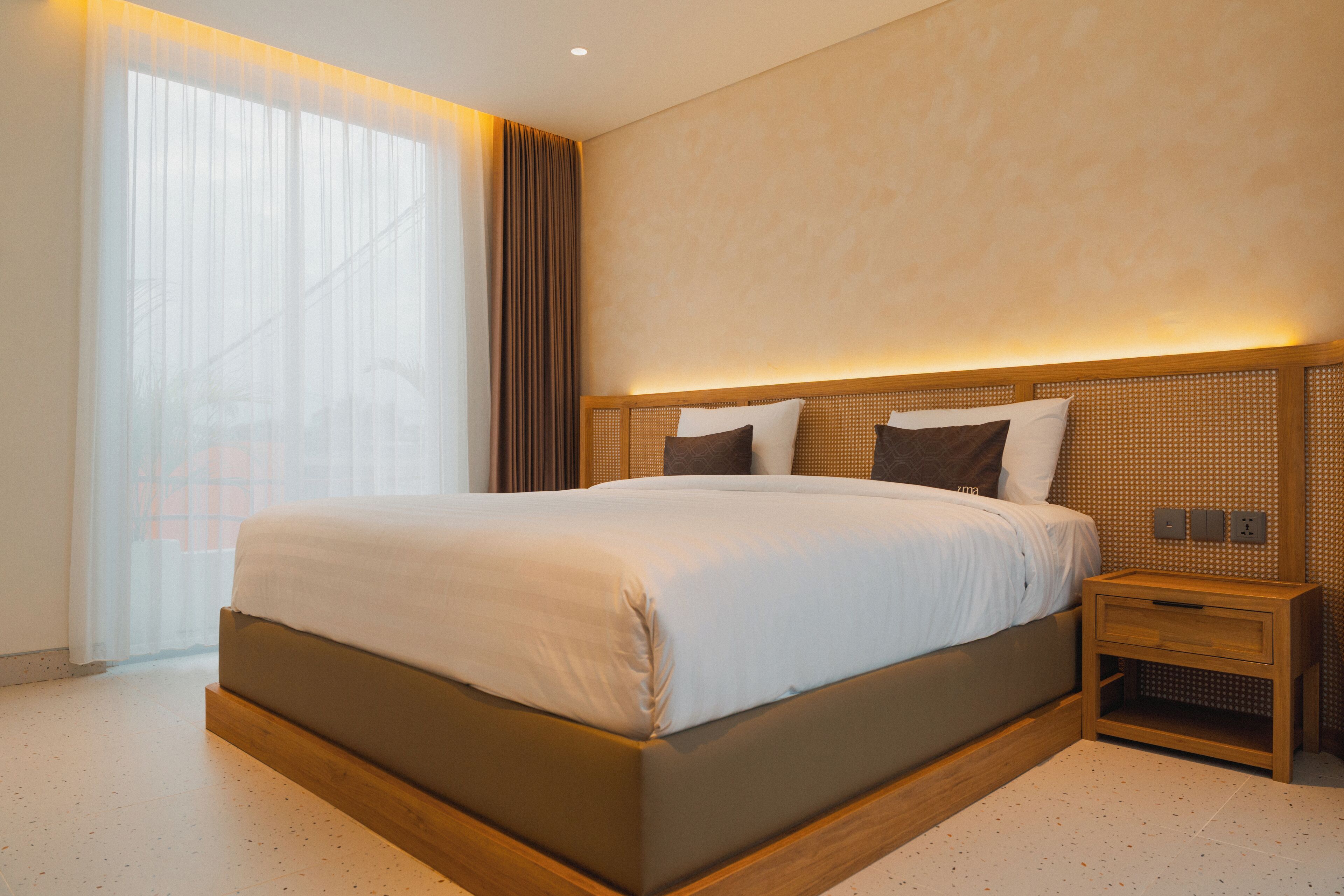 A modern hotel room with a large bed, a bedside table, and a window with sheer curtains.