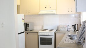 A compact kitchen with white appliances, including a refrigerator, microwave, stove, and toaster. There is a glass table and a chair in the foreground. - The Sundown Stay (Dubbo)