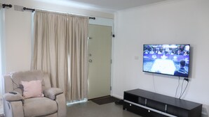 A living room with a beige armchair, a black entertainment unit, and a television displaying a sports event. - The Sundown Stay (Dubbo)