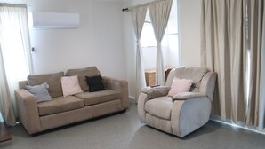 A living room with a beige sofa and armchair, beige curtains, and a window with curtains. - The Sundown Stay (Dubbo)
