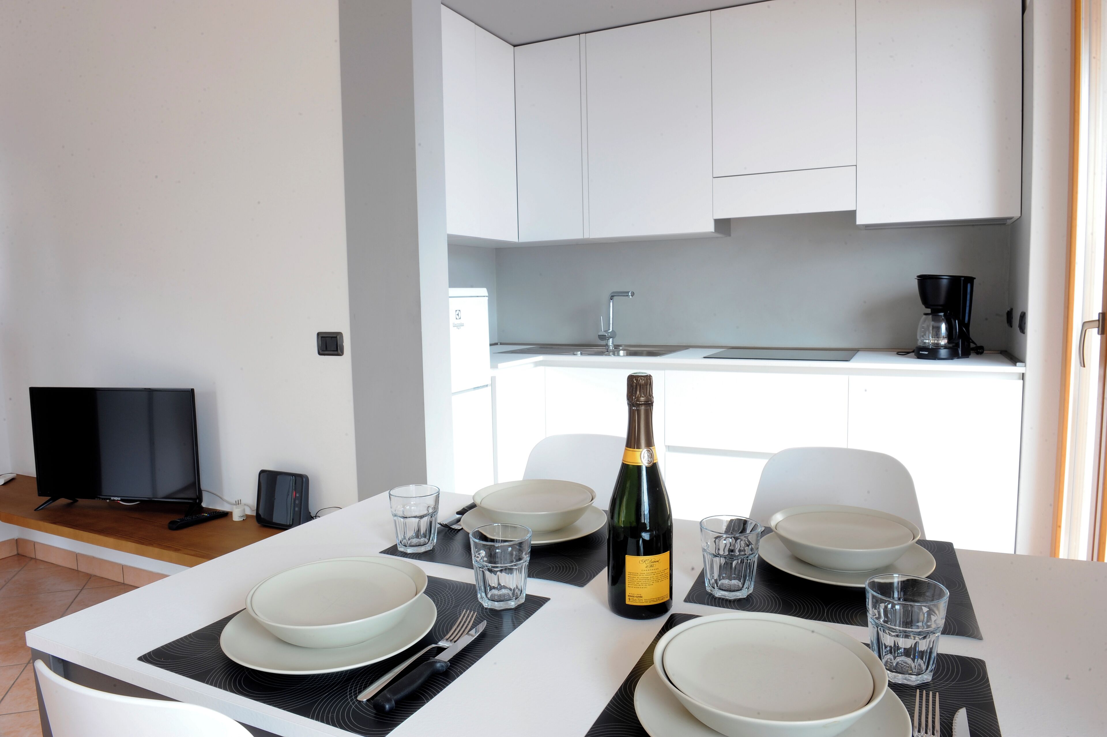 A modern kitchen with white cabinets, a black countertop, and a dining table set with plates, glasses, and a bottle of champagne.