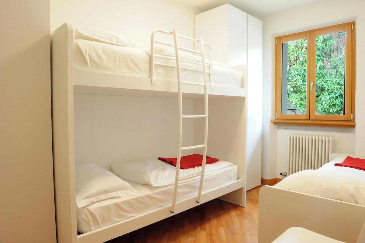 A bunk bed room with a ladder, a window with a view of greenery, and a radiator.