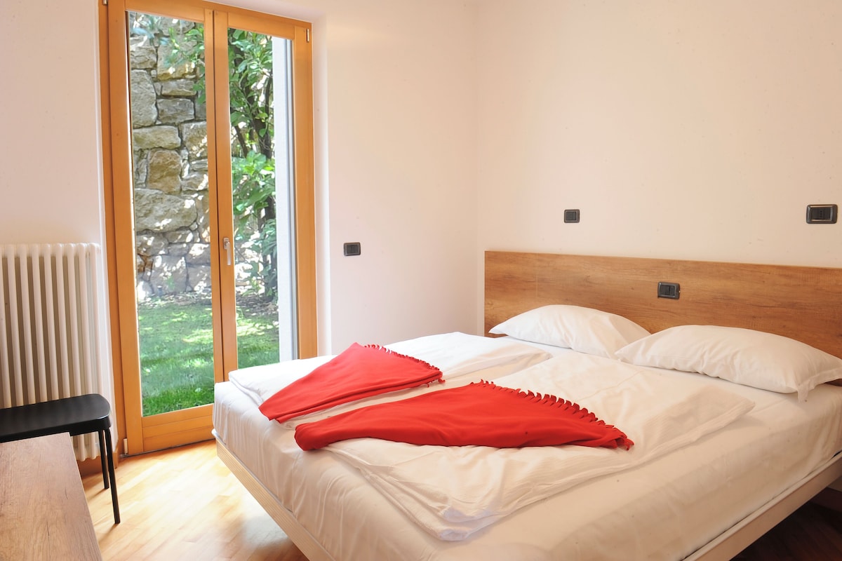 A bedroom with a wooden headboard, a large window with a stone wall view, and a red blanket on the bed.
